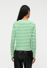 A hosszú ujjú póló zöld és fehér vízszintes csíkokkal rendelkezik, kerek nyakkivágású, és Karl Lagerfeld branding található a hátán. Fekete nadrággal párosítva.