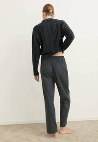 Maglione nero a trecce con design cropped e maniche lunghe; abbinato a pantaloni larghi grigi scuri con vita elasticizzata; piedi nudi visibili.