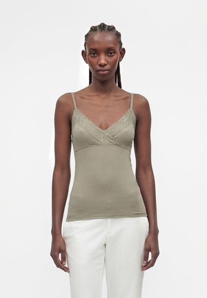 Femme aux cheveux tressés portant un caraco vert olive orné de dentelle et un pantalon blanc, debout devant un fond blanc uni.