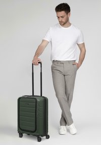 Valigia rigida verde con superficie texturizzata, zip nere, manico retractile e quattro ruote. Il modello si trova accanto ad essa.