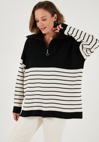 Schwarz-wei�ß gestreifter Pullover mit hohem Kragen und halbem Reißverschluss. Hergestellt aus weichem Strickmaterial, mit langen Ärmeln und lockerer Passform.