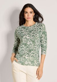 Eine Frau mit dunklem Haar trägt ein grünes und weißes, floral gemustertes Langarmoberteil und cremfarbene Hosen. Sie steht mit einer Hand in der Tasche vor einem neutralen Hintergrund.