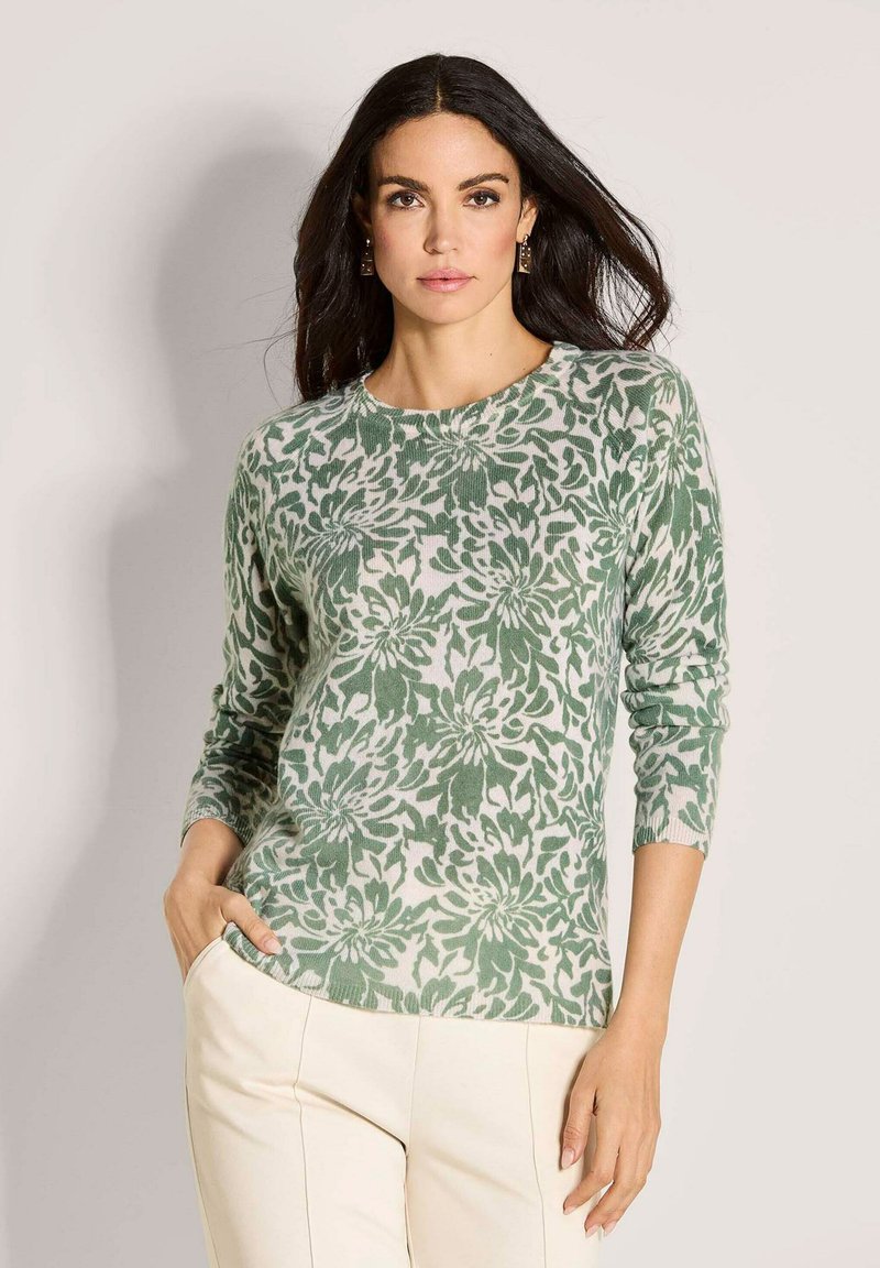 Eine Frau mit dunklem Haar trägt ein grünes und weißes, floral gemustertes Langarmoberteil und cremfarbene Hosen. Sie steht mit einer Hand in der Tasche vor einem neutralen Hintergrund.