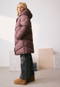 Kurtka puchowa w kolorze taupe z przeszyciami w jodełkę, w zestawieniu z szarymi spodniami w panterkę i beżowymi botkami za kostkę. W tle teksturowane bloki.