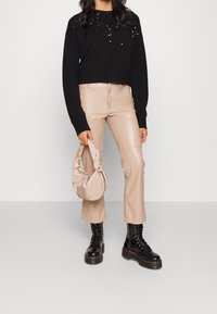 Czarny krótki sweter z cekinowym detalem, beżowe spodnie z ekologicznej skóry oraz czarne sznurowane buty. Trzyma beżową torbę zawiązaną na supeł.