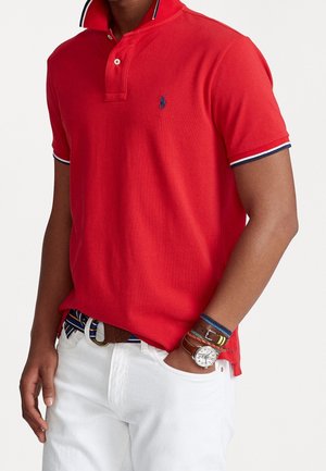 Rotes Poloshirt mit Kragen, kurzen Ärmeln und kontrastierendem Marineblau-weißem Besatz. Kombiniert mit weißen Hosen und mehreren Armbändern.