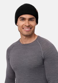 Beanie nero lavorato a maglia con texture a coste, dotato di un piccolo patch in pelle color noce. Indossato con una maglietta grigia a maniche lunghe aderente.
