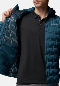 Turkusowa kurtka puchowa z poziomym pikowaniem, zapinana na zamek błyskawiczny, z czarnym wnętrzem. Noszona na czarnej koszulce polo z trzema guzikami.