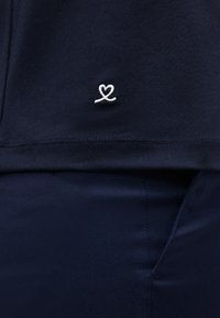 Ρουχισμός ναυτικού μπλε με μικρό λευκό κεντητό λογότυπο σε σχήμα καρδιάς πάνω από το ραμμένο στρίφωμα, σε συνδυασμό με αντίστοιχο ύφασμα ναυτικού μπλε από κάτω.