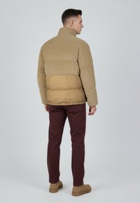 Kurtka z podszewką w beżowym kolorze, z teksturowaną górną częścią i gładką dolną. W zestawie z bordowymi spodniami i brązowymi butami. Wysoki kołnierz i prążkowany wzór.