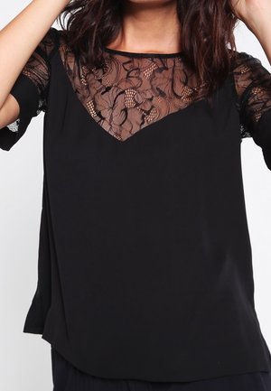 Schwarze Bluse mit transparentem floralen Spitzenbesatz über der oberen Brust und den Ärmeln, getragen von einer Person, die ihre Haare richtet.