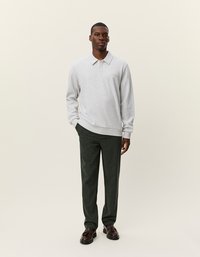 Szürke pulóver gallérral, sötétzöld nadrággal és barna cipőkkel párosítva. A modell világos háttér előtt áll.