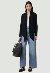 Cardigan preto de malha com frente aberta, combinado com calças largas de denim claro de cintura alta. Segura uma mala tote preta e usa sapatos planos pretos.