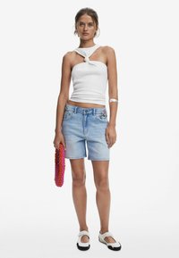 Vit knuten tanktop, ljusblå denim shorts med blommönster, håller en färgglad pärlryggsäck. Svart-och-vit slip-on skor.