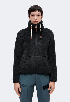 Persona que lleva una chaqueta de forro polar negra con un cuello alto y cordones, combinada con pantalones azul grisáceo con detalles de parches negros.