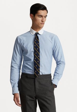 Cămașă lungă albastru deschis cu guler alb, asortată cu o cravată albastru închis cu dungi aurii și modele de embleme. Pantaloni gri completează ținuta.