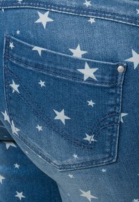 Jean en denim avec un motif étoilé blanc sur fond bleu. Poche arrière visible avec des détails de couture et un rivet argenté.