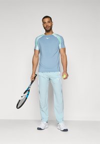 Lys blå treningsskjorte med mintgrønne detaljer, paret med matchende lys blå bukser og hvite joggesko. Holder en tenniskøll og ball.