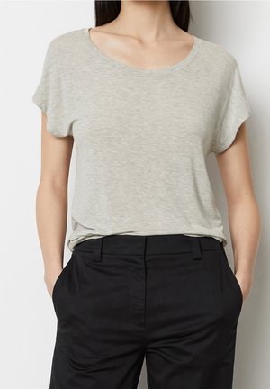 Frau trägt ein hellgraues T-Shirt mit kurzen Ärmeln und rundem Ausschnitt, das in eine hoch taillierte schwarze Hose gesteckt ist, mit den Händen in den Taschen.