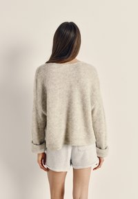 Gaiši pelēks, pārmēroga džemperis no mīksta, teksturēta adījuma ar aizlocītām aprocēm. Kombinēts ar gaiši pelēniem ikdienišķiem šortiem. Skats no aizmugures uz neitrālas fona.