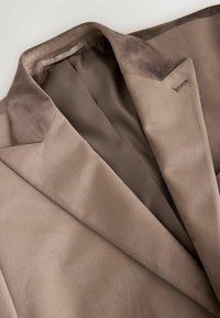 Hellbraune Jacke mit einer glatten, wildlederähnlichen Textur und einem kontrastierenden dunkleren Kragen. Das Innere verfügt über ein Mesh-Futter für Atmungsaktivität.