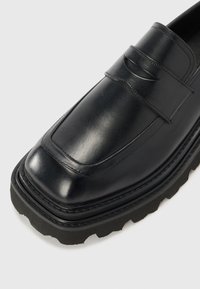 Svart läderloafersko med rektangulärt tåskydd, dekorspänne och kontrasterande tjock gummisula med en strukturerad vågmönster.