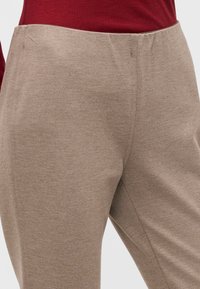 Gros plan sur un pantalon beige en maille porté avec un haut côtelé rouge, montrant un tissu lisse et une taille plate et élastique.