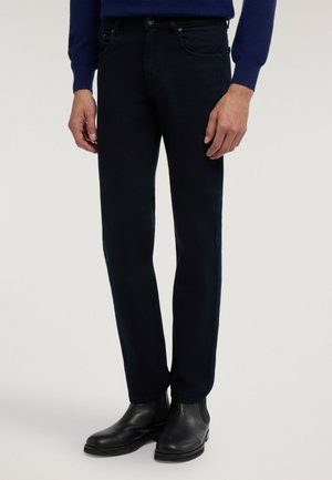 Man som bär mörka slim fit-byxor med svarta läderstövlar och en mörkblå långärmad topp, står mot en enfärgad bakgrund.