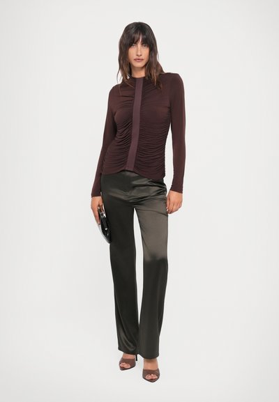 Brown long-sleeve top with textured ruching and front panel detail, paired with dark olive satin trousers and a black clutch bag.