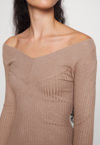 Femme portant un pull beige côtelé, épaules découvertes, montrant le cou et les épaules, avec de longs cheveux bouclés foncés visibles derrière.