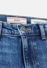 Close-up van de tailleband van blauwe spijkerbroeken met metalen knoop en zichtbare "GUESS" merklabel en "OVERSIZE FIT" tag aan de binnenkant.