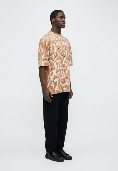 Άνδρας που φοράει καφέ μπλουζάκι Moschino με σχέδια, μαύρο παντελόνι και μαύρα αθλητικά παπούτσια σε απλό λευκό φόντο.