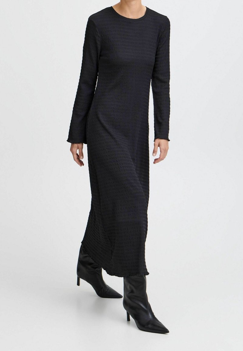Femme portant une robe maxi noire texturée à manches longues, associée à des bottines à talons noires à bout pointu, marchant devant un fond uni.