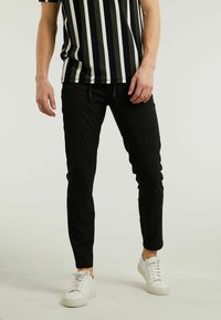 Zwarte tapered broek met een drawstring taille, gecombineerd met een zwart-wit gestreept shirt. Model draagt witte sneakers. Effen achtergrond.