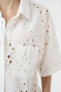 Camicia bianca ricamata caratterizzata da un motivo floreale texturizzato, maniche corte, chiusura a bottoni e una tasca sul petto con dettagli ritagliati.