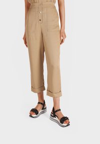 Pantaloni beige a vita alta con una vestibilità ampia, dotati di tasche anteriori e orli arrotolati, abbinati a sandali con zeppa neri con strisce bianche.