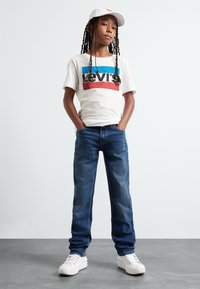 White t-shirt with red and blue Levi's logo, blue denim jeans, and white sneakers. The person has braided hair, wearing a white cap.
