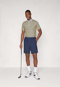 Polo tričko s krátkým rukávem s vzorem v žluté a modré barvě, spárované s námořnickými šortkami. Bílé sportovní boty a golfová hůl v ruce.