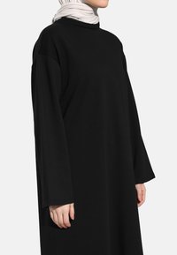 Long black dress with a round neck and wide sleeves, made of soft fabric. Model wears a light-colored textured hijab.