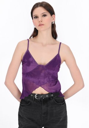 Giovane donna con capelli castani lisci, che indossa orecchini d’argento, un top viola trasparente senza maniche e jeans neri a vita alta con cintura.