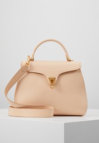 Beige texturerad läderhandväska med ovandelshandtag, justerbar axelrem och klädd i guldton, visad på en vit pelare mot en enkel bakgrund.