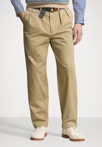 Man med beige veckade byxor, randig blå skjorta, mönstrad stickad väst och vita snörskor, stående med händerna i fickorna.