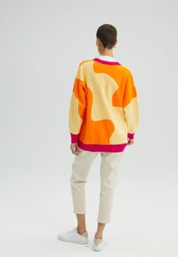 Sweter z dzianiny w kolorze pomarańczowym i kremowym z różowymi akcentami, zdobiony abstrakcyjnymi wzorami. Połączony z beżowymi spodniami i białymi tenisówkami.