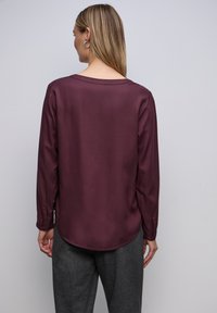 Langärmliges Shirt aus tiefburgunder Samt mit rundem Halsausschnitt und abgerundetem Saum. Mit Knopfmanschetten und glatter Textur. Getragen mit grauen Hosen.