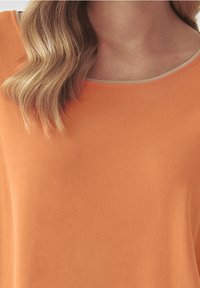 Oranžový top s krátkými rukávy a hladkým povrchem, s kulatým výstřihem a tenkým kontrastním lemem kolem krku.