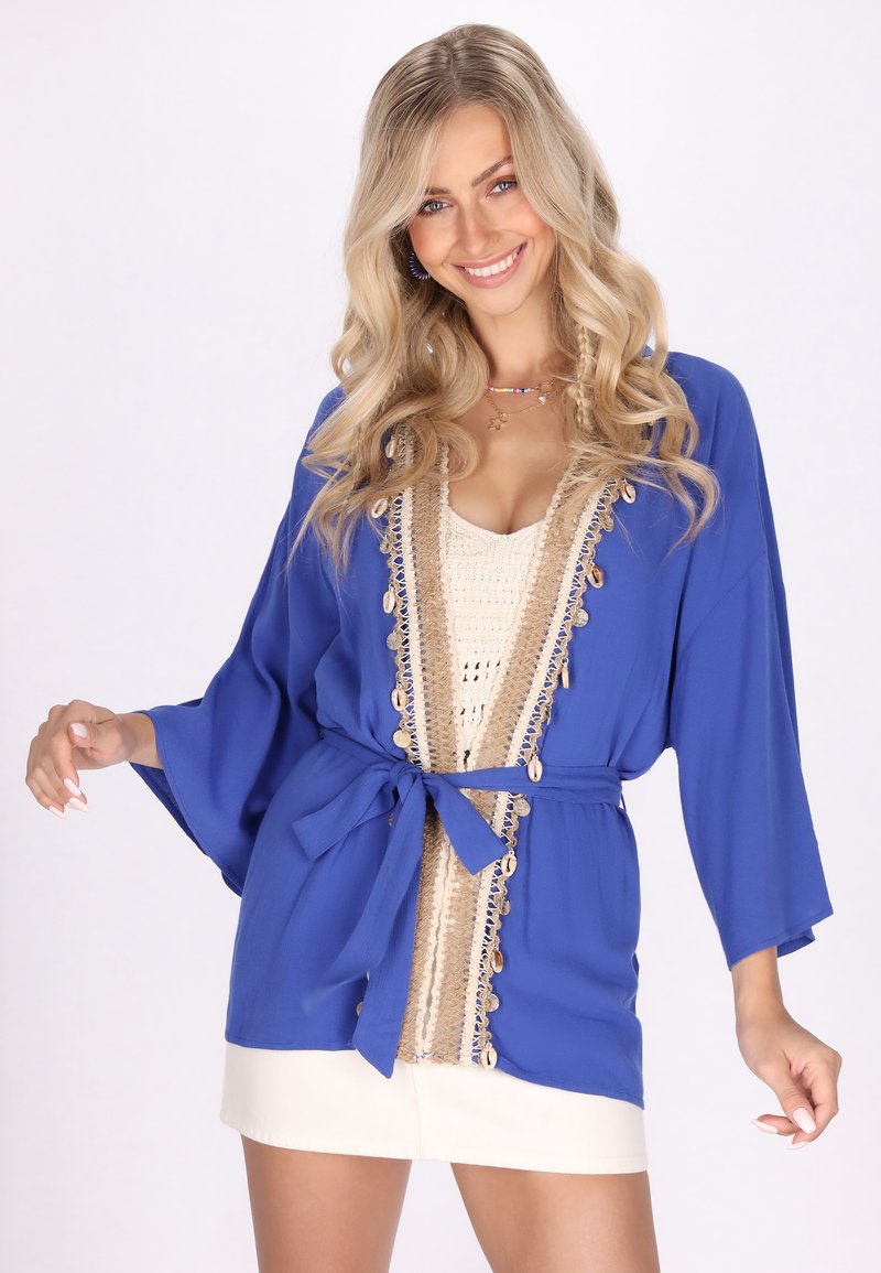 Plavi kimono stil jakna s širokim rukavima, bež obrubom i školjkastim akcentima, te vezanjem u struku. Lagana tkanina s glatkom teksturom.
