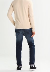 Beiger Pullover mit langen Ärmeln, kombiniert mit blauen Jeans aus Denim mit einem dezenten Ausbleichungseffekt. Schwarze Sneaker runden das Outfit ab.