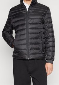 Muž oblečený v černé prošívané bundě na zip s bočními kapsami a elastickými manžetami, kombinované s černými kalhotami a bílou košilí pod tím.