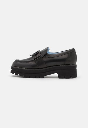 Sort læder loafer med chunky sål, sidestrop med spænde og lav hæl på en plain lys baggrund.