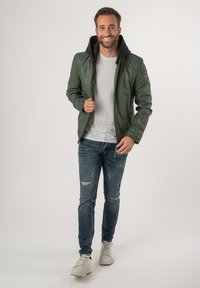 Giacca in pelle verde con cappuccio nero, abbinata a una maglietta bianca e jeans blu strappati. Sneakers bianche completano l'outfit.